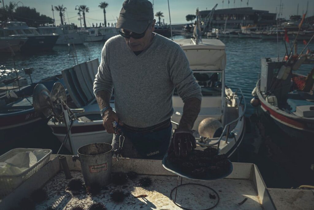 A mechanic working on a center console boat tied at a marina slip, checking the engine compartment with open hatch lids, a portable diagnostic kit, tools laid out on the dock, and nearby pilings and calm water in the background