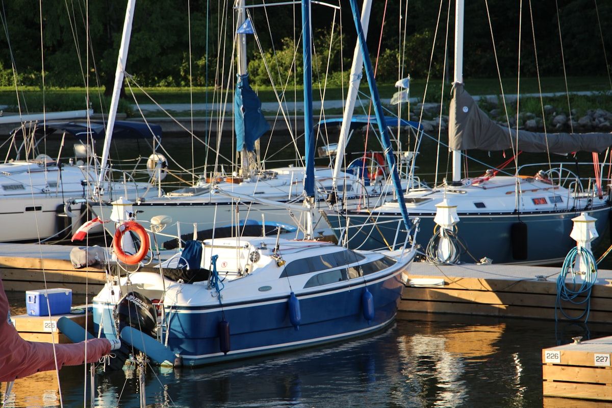 A small recreational boat tied to a marina slip beside cleaning supplies, coiled dock lines, a battery charger, a service bucket, and a pair of work gloves, with a cluttered dock showing signs of multiple pending maintenance tasks