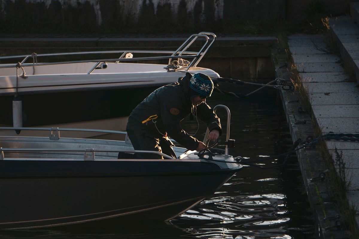 A marine mechanic standing on a dock beside a powerboat, using a toolbox and handheld diagnostic gear while the boat remains tied up in its slip, with marina water, pilings, and nearby lifts in the background