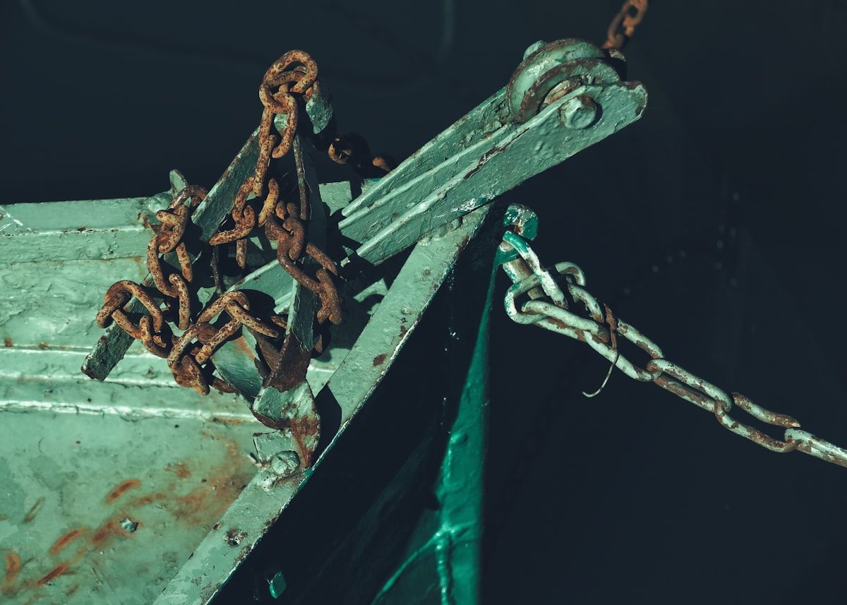 A salt-weathered boat engine bay showing corroded metal fittings, battery terminals, hoses, and electrical connections, with bright sunlight reflecting off humid marina surroundings and a dockside service cart nearby