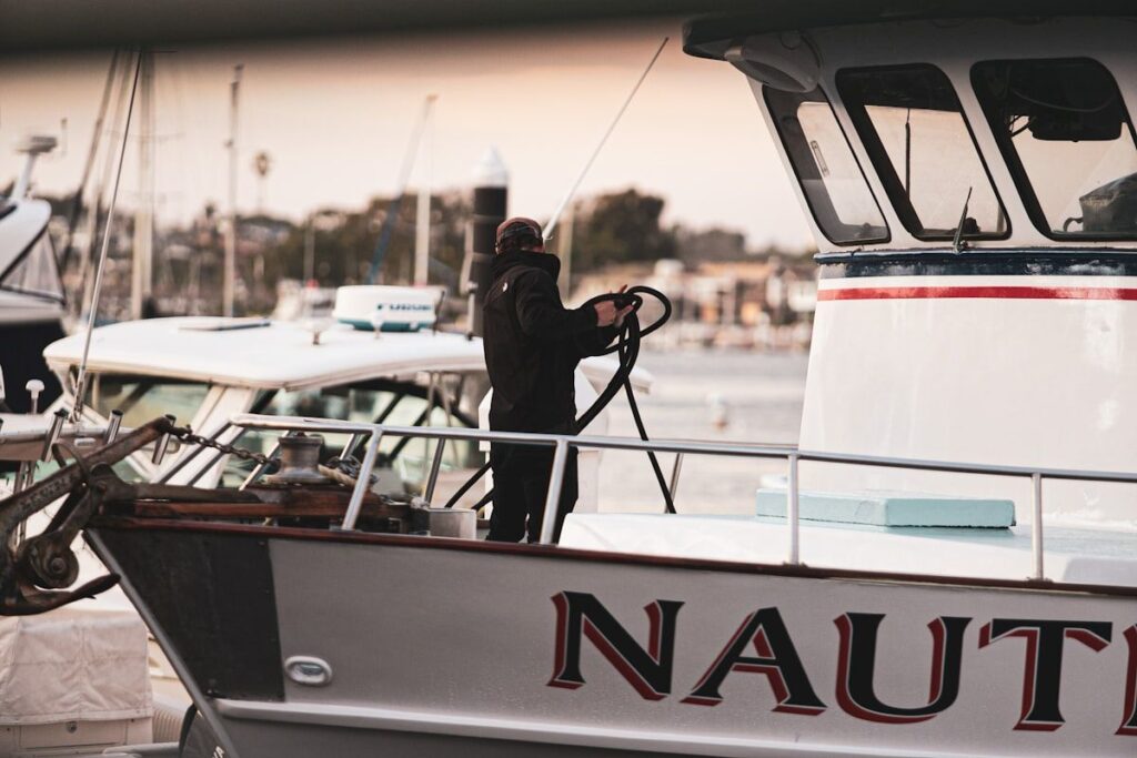 A boat at the dock being serviced with a pressure washer on the hull, a detailer wiping down stainless railings and vinyl seats, and an open engine hatch showing a mechanic changing filters and inspecting belts and fluid reservoirs.