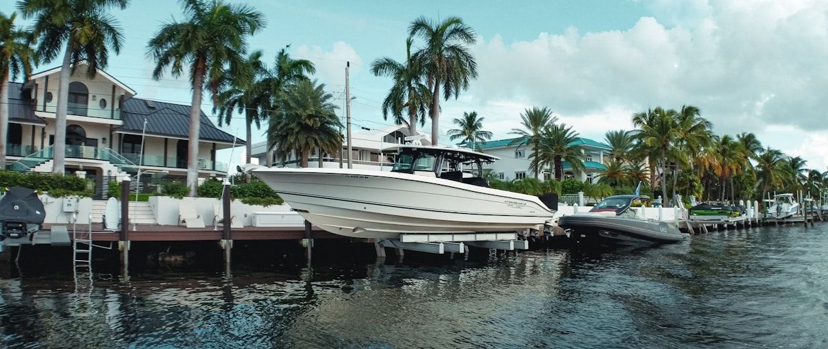 A white fiberglass boat tied at a southwest Florida marina with green saltwater staining near the waterline, barnacle growth on the lower hull, a worn zinc anode on the trim tab, sun-faded seat cushions under a canvas cover, and storm-prep lines and fenders secured along the dock.