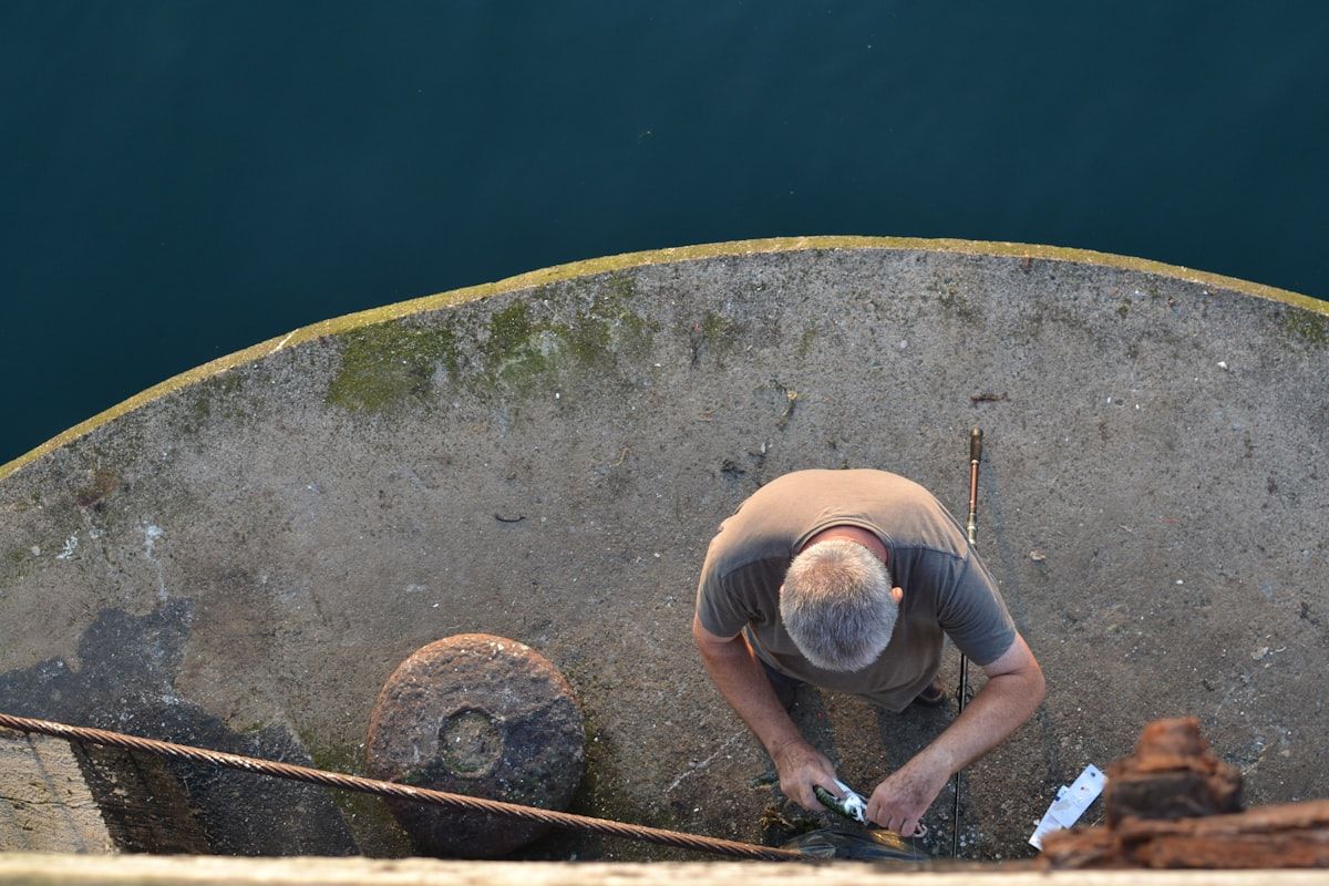 A marine technician standing on a dock beside a moored center-console boat, inspecting the engine compartment with tools laid out on the dock, with the boat still tied in its slip at a marina