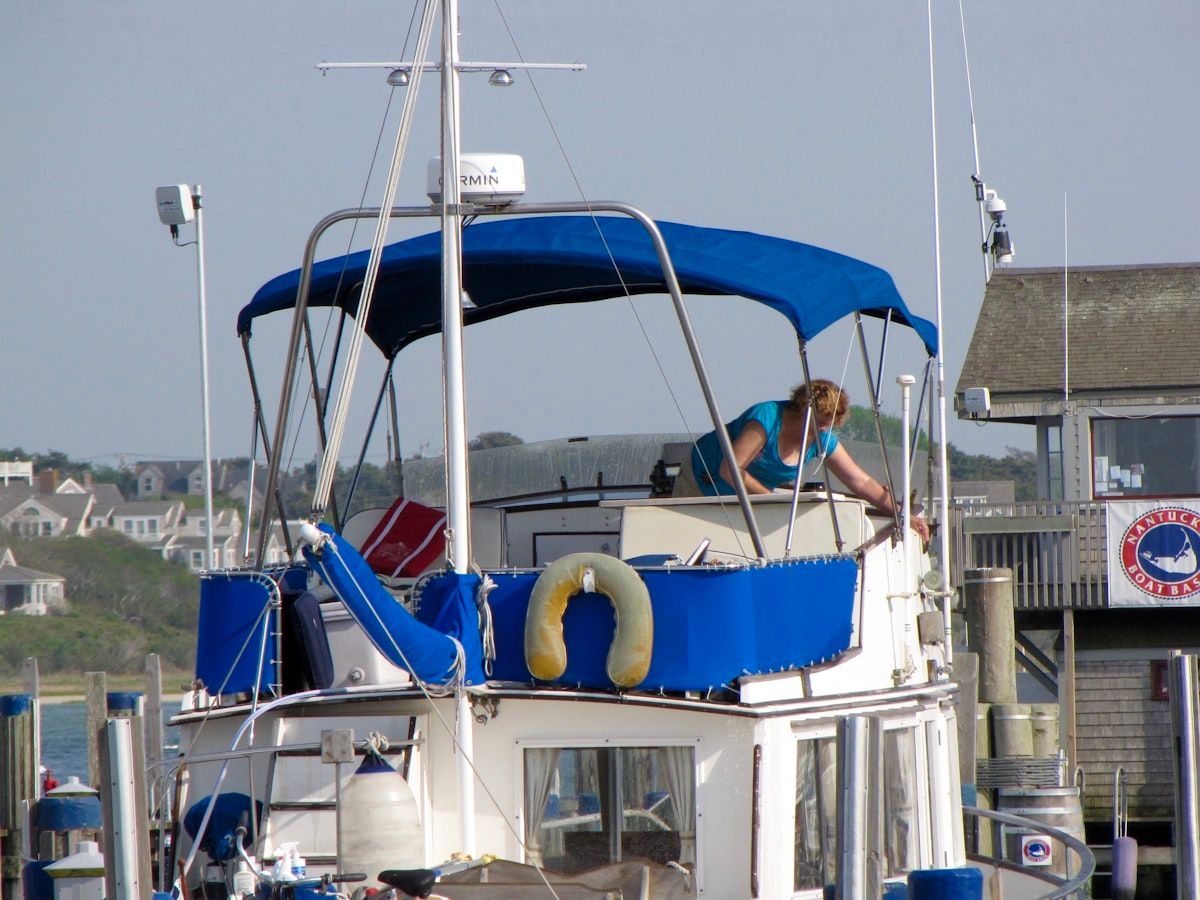A sportfishing boat in a marina slip with the engine hatch open, a marine technician tracing corroded wiring near a trim-tab control and checking an exposed battery compartment while tools and salt-stained hoses sit on the deck